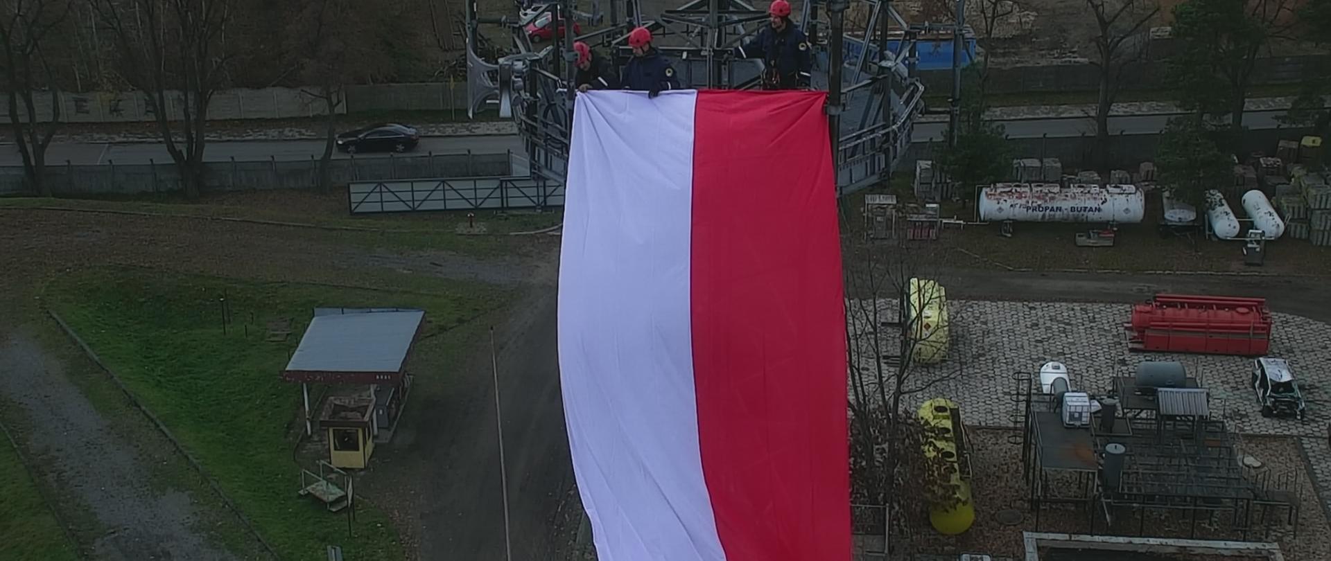 Na zdjęciu troje funkcjonariuszy sekcji wysokościowej CS PSP wiesza biało-czerwoną flagę na platformie masztu kratownicowego