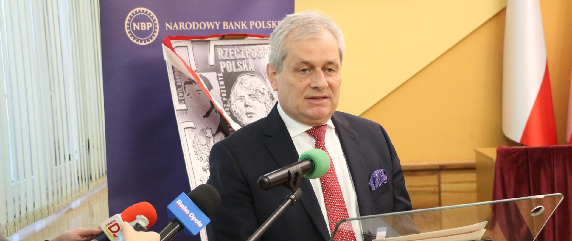 Na zdjęciu wojewoda opolski, który stoi na przeciw dziennikarzy. Przed nim stoi szklana mównica. Wojewoda opiera dłonie o mównice. Po lewej stronie widoczni dziennikarze, trzymający mikrofony. W tle baner NBP.