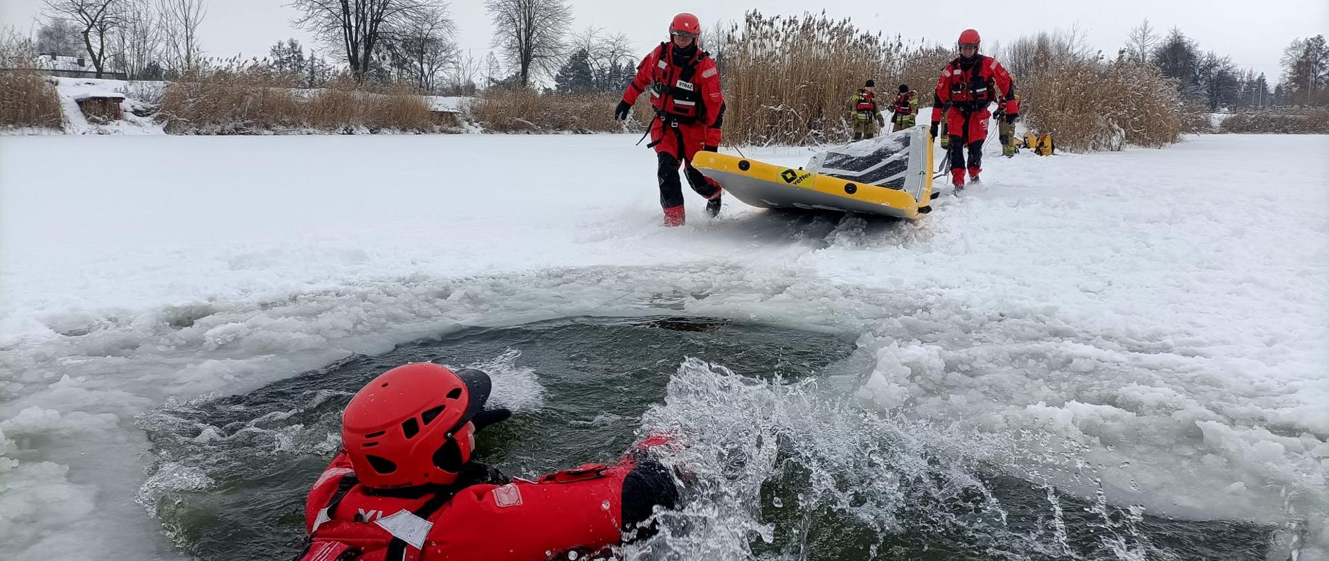 Na zamarzniętym zbiorniku wodnym, W przeręblu znajduje się ratownik. Po lodzie biegnie do niego dwóch innych, trzymając żółty ponton lodowy. W tle wzniesienie zarośnięte krzakami, a dalej drzewa bez liści. Powyżej pochmurne niebo. 