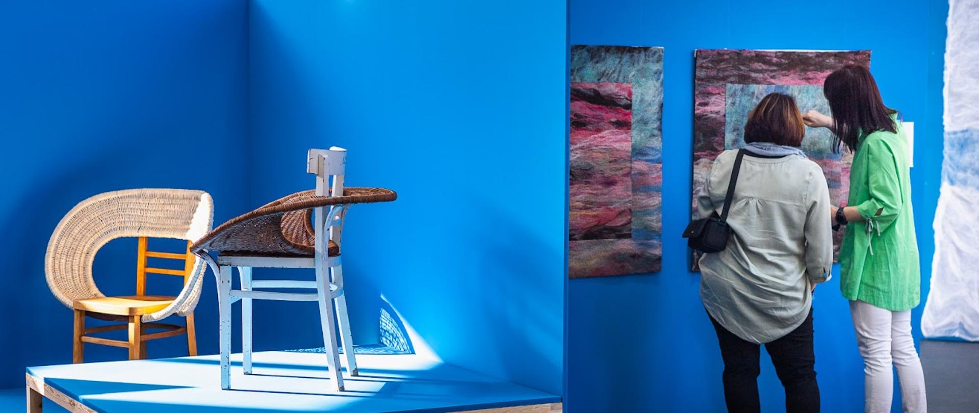 Zdjęcie wykonane podczas wystawy prac na Biennale 2022 w PLSP w Nałęczowie. Przedstawia prace uczniów, które są wyeksponowane przy lu na niebieskich ścianach. Po lewej stronie zdjęcia na podestach stoją 2 krzesła, po prawej przy pracach wiszących na ścianie stoi 2 oglądające je osoby.