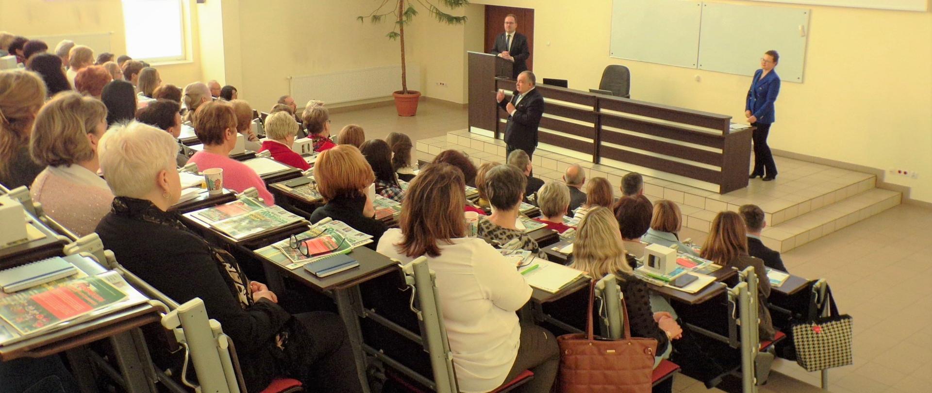 Na sali wykładowej duża grupa osób, głównie kobiet, słucha wystąpienia dyrektora OR, za którym stoi duże biurko – katedra. Po lewej stronie przy mównicy stoi mężczyzna, po prawej stronie biurka stoi kobieta.