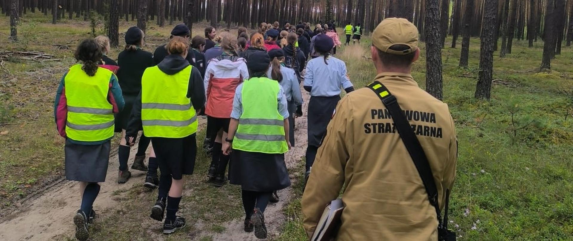 Fotografia przedstawia próbną ewakuację uczestników obozu pod nadzorem funkcjonariusza Państwowej Straży Pożarnej. Leśną drogą idzie grupa młodzieży, którą zamykają 3 osoby kadry obozu i strażak. 