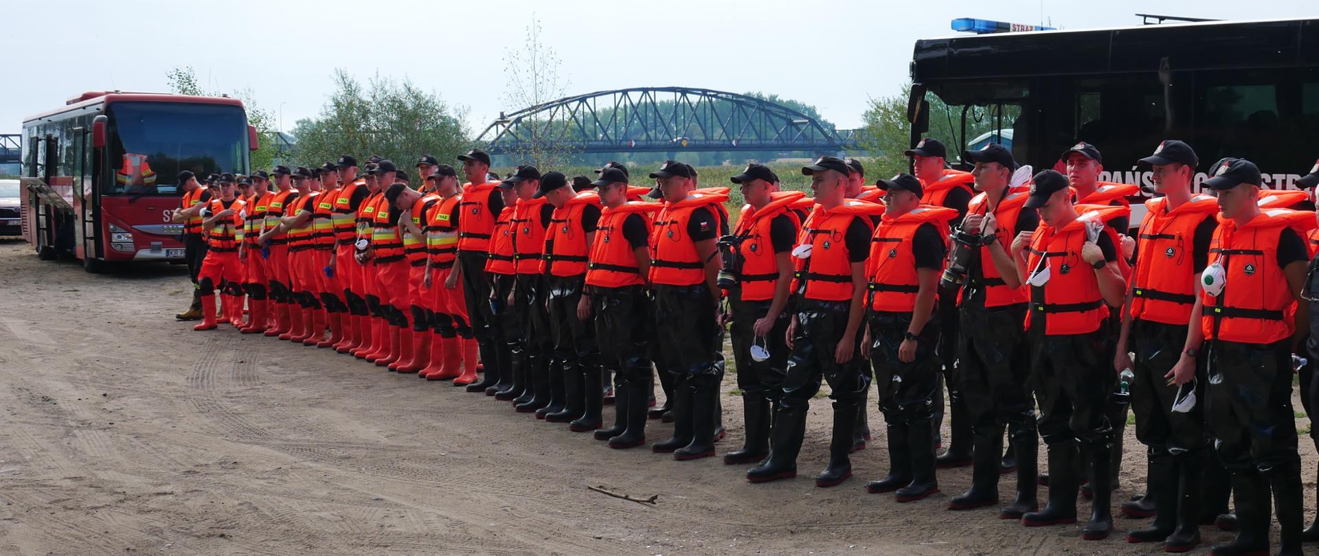 Grupa kilkudziesięciu strażaków stojąca w dwuszeregu w trakcie odprawy przed działaniami. Część z nich w ubraniach w kolorze pomarańczowym pozostali w ubraniu koszarowym w kolorze czarnym. W tle autobusy strażackie. 
