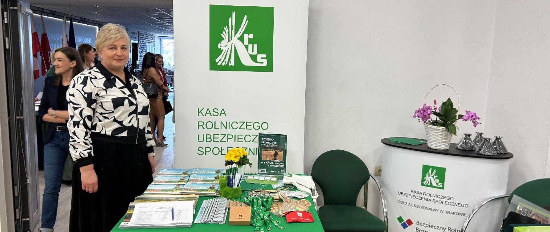 Kobieta stoi przy stole nakrytym obrusem z logo KRUS, na którym leżą ulotki i foldery obok stojak reklamowy z logo KRUS.