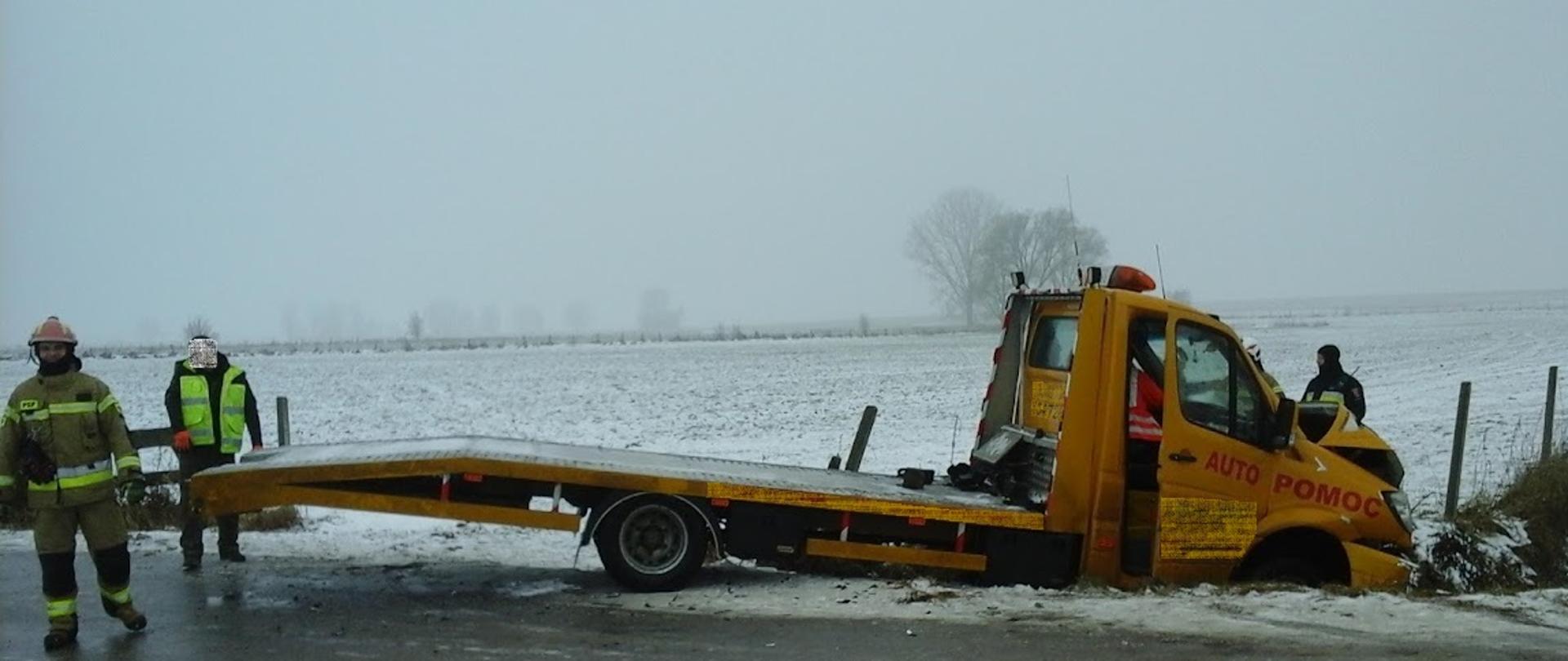 Samochód ciężarowy - autolaweta, stojący przodem w przydrożnym rowie, tył pozostaje na jezdni. Przód samochodu uszkodzony.