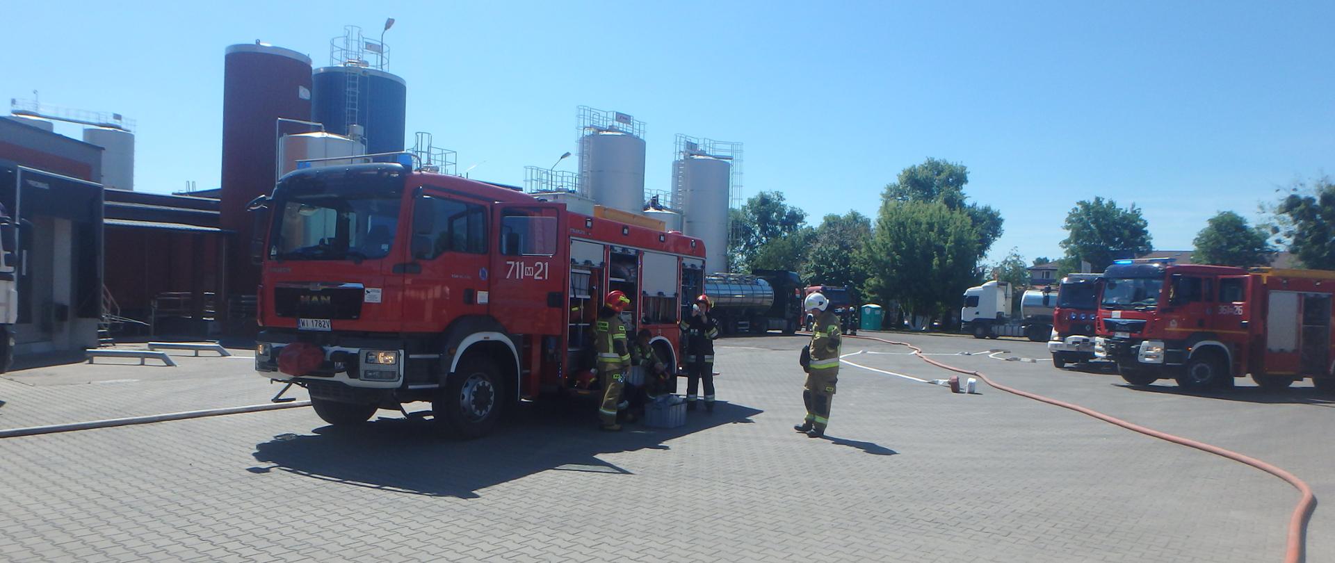 Samochody strażackie stoją na terenie mleczarni. Obok stoją strażacy z OSP i PSP przygotowując się do odbycia ćwiczeń.