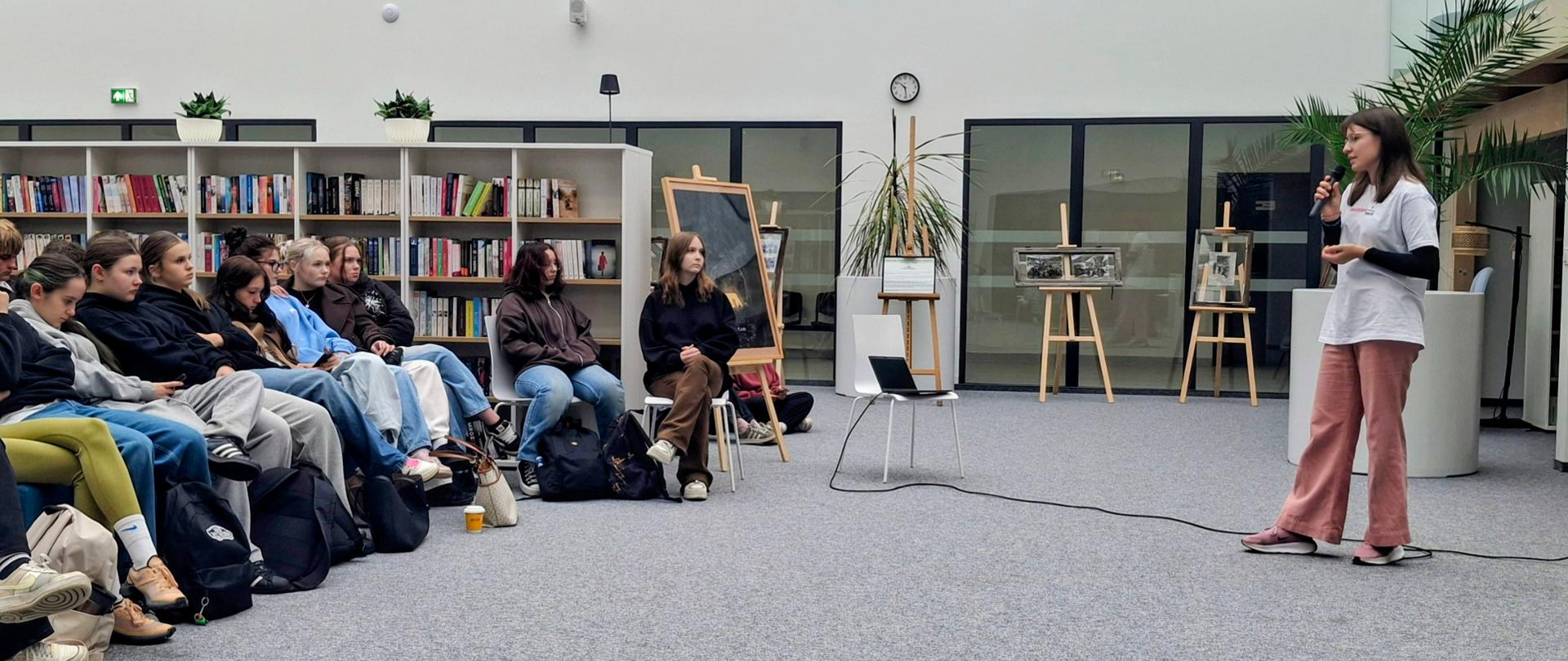 Zdjęcie przedstawiające kobietę przemawiającą do uczniów w bibliotece szkolnej