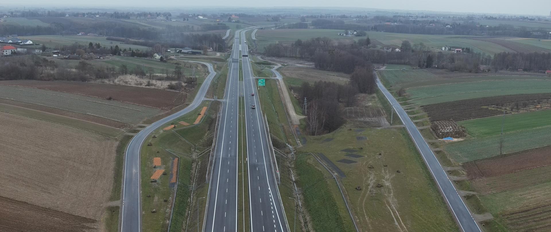 obwodnica Opatowa - ujęcie z góry, centralnie przez środek zdjęcia dwujezdniowa asfaltowa droga, po obu stronach tej trasy biegną wzdłuż niej jezdnojezdniowe drogi dojazdowe, przy drogach pola, w oddali zarośla i zabudowania 