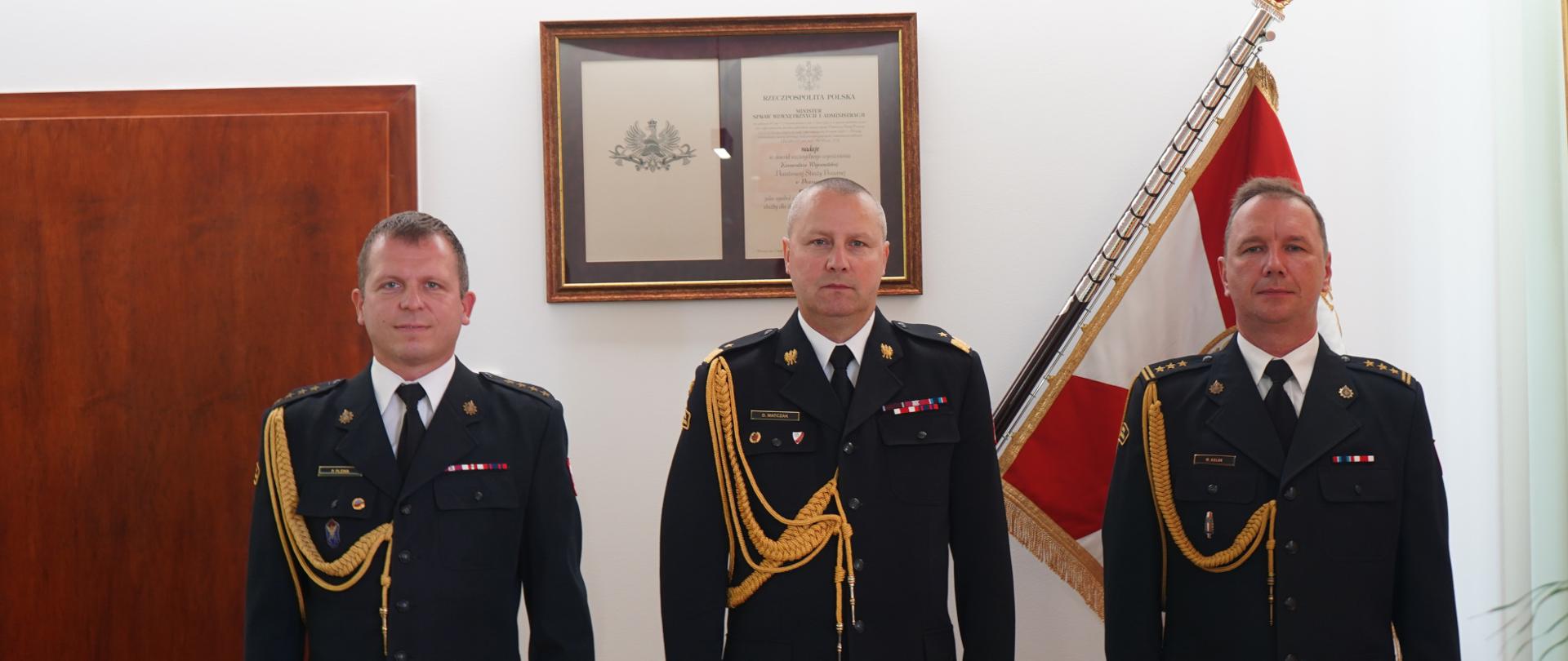 Zdjęcie przedstawia trzech funkcjonariuszy Państwowej straży Pożarnej w mundurach wyjściowych ze sznurem galowym. W środku Komendant Wojewódzki PSP po obu stronach funkcjonariusze, którzy odbierali akty powołania na kierownicze stanowisko w jednostkach organizacyjnych PSP