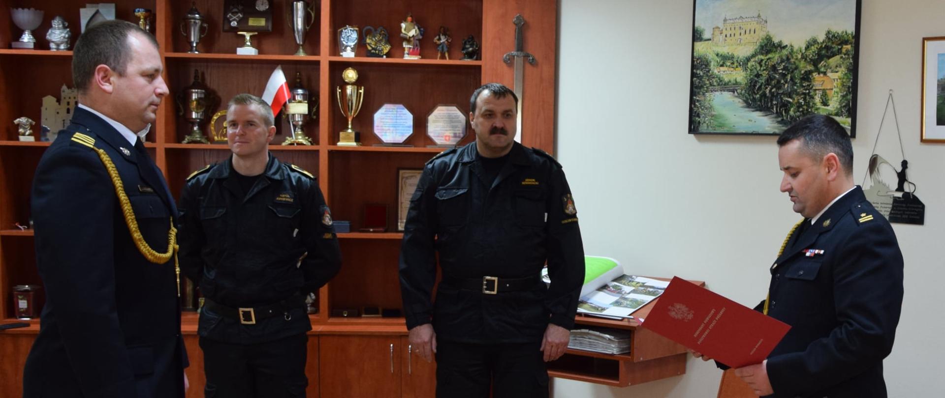 Dwóch strażaków w umundurowaniu galowym stoi na przeciwko siebie, Strażak po lewej stronie trzyma w dłoniach bordową teczkę, W tle widać dwóch strażaków w umundurowaniu dowódczo-sztabowym.