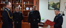 Dwóch strażaków w umundurowaniu galowym stoi na przeciwko siebie, Strażak po lewej stronie trzyma w dłoniach bordową teczkę, W tle widać dwóch strażaków w umundurowaniu dowódczo-sztabowym.