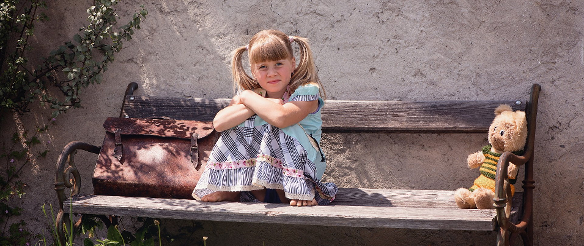 dziewczynka w letniej sukience siedząca na starej ławce pod wiejskim domem, po jej lewej stronie leży walizka/tornister, po prawej siedzi misiek.
