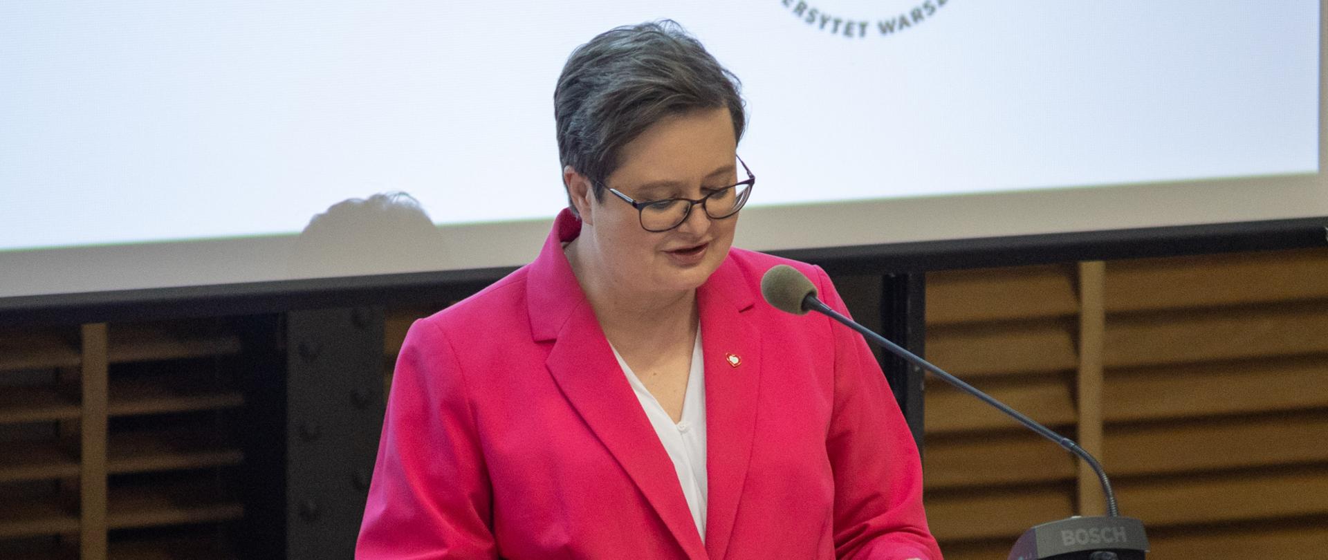 Wiceminister Katarzyna Lubnauer, ubrana w różowy garnitur i białą bluzkę, z przypinką w klapie biało-czerwonego serca, przemawia z mównicy, na której widnieje herb Uniwersytetu Warszawskiego. Na mównicy stoi czarny mikrofon. W tle widać planszę ze szczegółami dotyczącymi Olimpiady.
