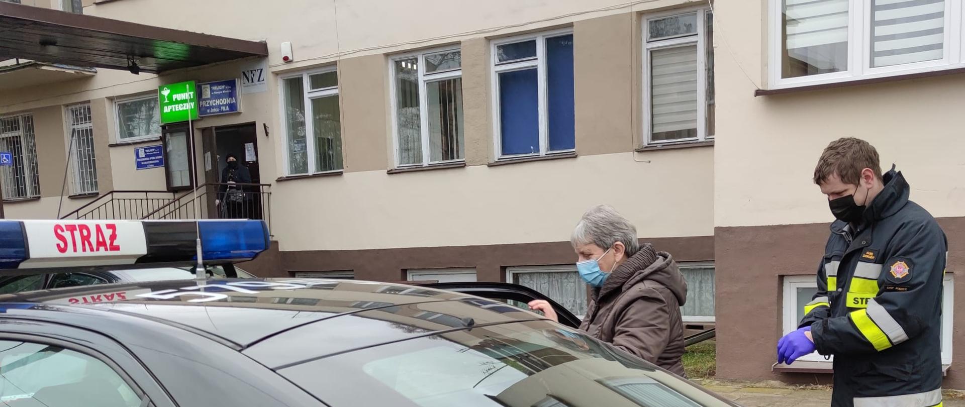 Na pierwszym planie fragment samochodu pożarniczego osobowego w kolorze ciemnym z czerwonym pasem i białym napisem OSP Joniec. Za samochodem strażak w ubraniu koszarowym, obok przewożona osoba.