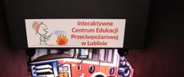 Grupa strażaków PSP w mundurach schodzi po schodach do Interaktywnego Centrum Edukacji Przeciwpożarowej w Lublinie, nad nimi szyld z nazwą placówki i rysunkiem dziecka gaszącego ogień.