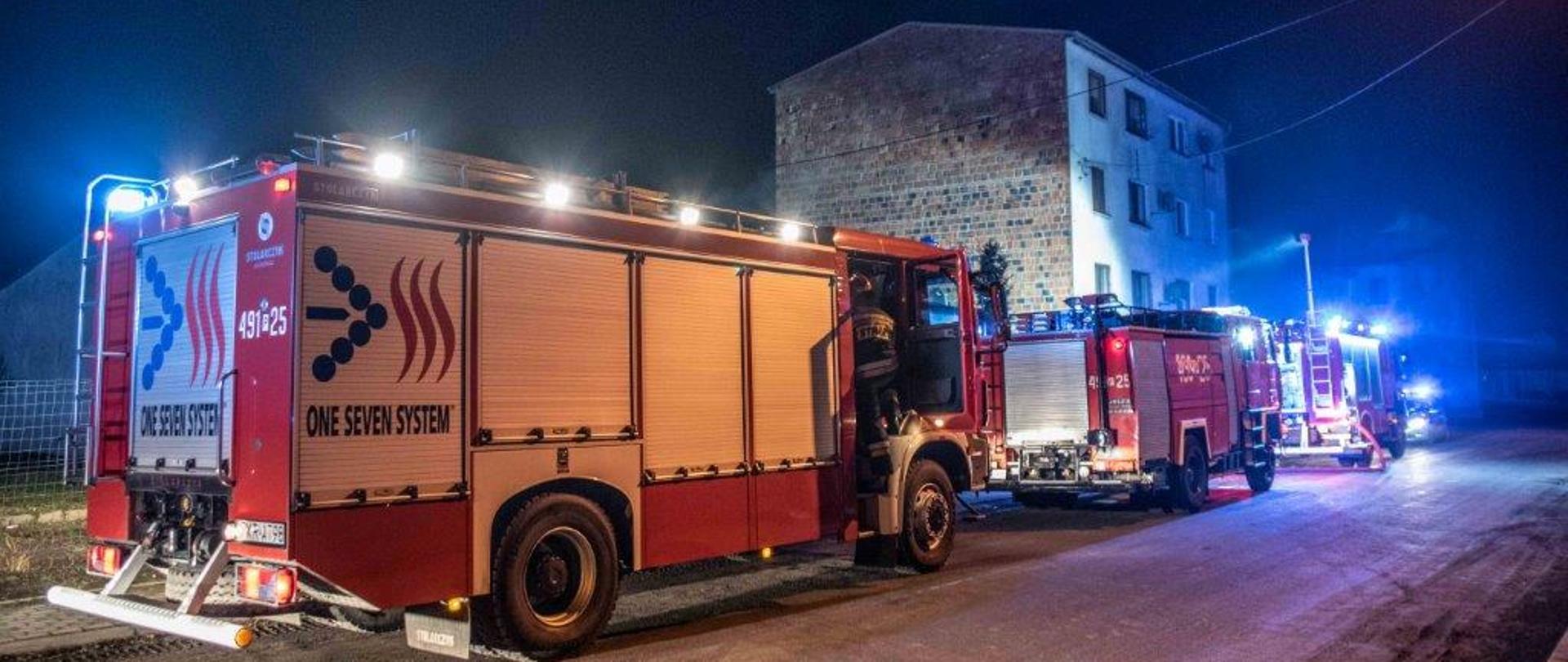 Trzy samochody gaśnicze ustawione w kolumnie na drodze przed budynkiem. Pierwszy pojazd oświetla teren działań masztem oświetleniowym. Pojazdy mają włączone sygnały błyskowe. W tle trzypiętrowy budynek objęty działaniami. 