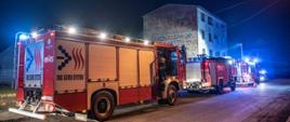 Trzy samochody gaśnicze ustawione w kolumnie na drodze przed budynkiem. Pierwszy pojazd oświetla teren działań masztem oświetleniowym. Pojazdy mają włączone sygnały błyskowe. W tle trzypiętrowy budynek objęty działaniami. 