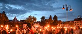 Tłum mieszkańców w różnym wieku, w tym rodziny z dziećmi i wózkami, spaceruje po wybrukowanym placu, niosąc zapalone pochodnie, które rozświetlają scenę ciepłym blaskiem. W tle widoczna jest sylwetka zabytkowej karuzeli oraz zarysy kamienic i drzew na tle ciemniejącego, zachmurzonego nieba.