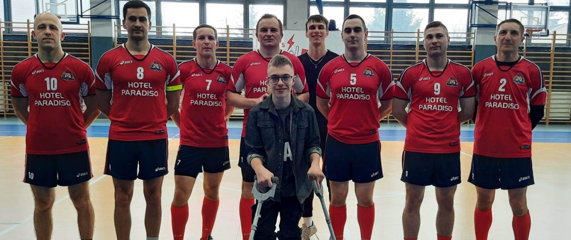 Strażacy z KP PSP Skarżysko-Kamienna pozują z chorym Piotrem do zdjęcia podczas turnieju charytatywnego. Strażacy są ubrani w stroje sportowe. Zdjęcie wykonano w hali sportowej.