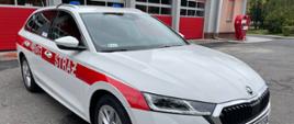 NOWY SAMOCHÓD W KOŚCIAŃSKIEJ KOMENDZIE. Na zdjęciu samochód Skoda Octavia, kolor biały, czerwony pas na wysokości klamek od drzwi z napisem straż i numer operacyjny, w tle budynek Komendy. 
