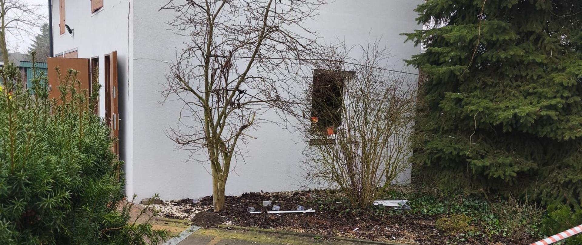 Fragment posesji z jasnym budynkiem gospodarczym widzianym z boku, przed którym znajduje się niewielki parking, oddzielony biało-czerwoną taśmą. Przy ścianie rosną bezlistne krzewy i małe drzewo, a po prawej stronie widoczna jest wysoka choinka; na ziemi leżą drobne fragmenty i zanieczyszczenia po wybuchu, który miał miejsce w budynku.