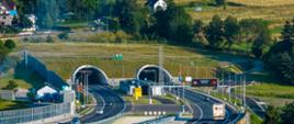 Widok na dwunawowy wylot tunelu drogowego położonego w pagórkowatym, zielonym terenie; przed portalami biegnie wielopasmowa droga ekspresowa z ekranami akustycznymi i tablicami zmiennej treści.