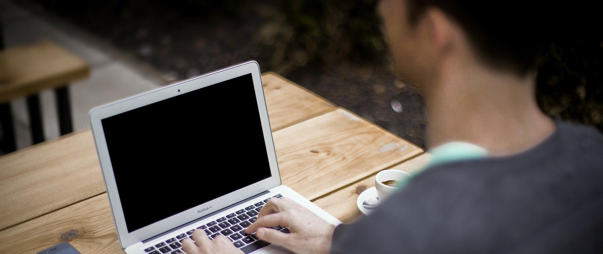 Na zdjęciu mężczyzna siedzi przy drewniany stole i pisze na laptopie. Na stole obok laptopa leży telefon komórkowy i notes z długopisem, obok stoi filiżanka z kawą.
