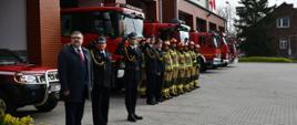 Zdjęcie przedstawia strażaków, którzy stoją na placu przed garażami oraz starostę powiatu bocheńskiego z okazji zbiórki dotyczącej podniesienia flagi państwowej. Część strażaków ubrana jest w stroje galowe, a część w stroje bojowe. Strażacy i Pan Starosta mają wzrok skierowany na podnoszoną flagę.