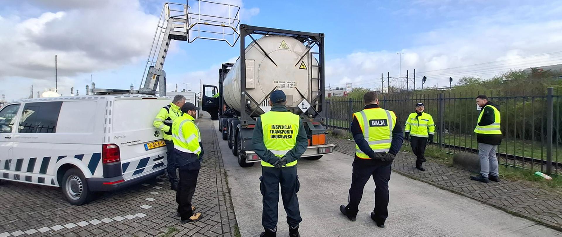 Grupa inspektorów transportu drogowego stoi wokół cysterny chemicznej podczas kontroli na terenie przemysłowym.