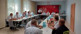Zdjęcie przedstawia druhów siedzących przy stole na zebraniu. Przy środkowym stole stoi Komendant Miejski PSP i wita gości. W tle ścianka z godłem.