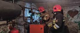 Na pierwszym planie dwóch strażaków sprawdzających przewód kominowy w kotłowni. 