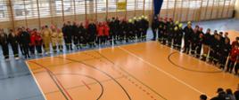 Zawodnicy startujących drużyn stoją grupowo na sali po wykonanych konkurencjach, stoją na kształcie kwadratu, oczekują na wyniki zawodów