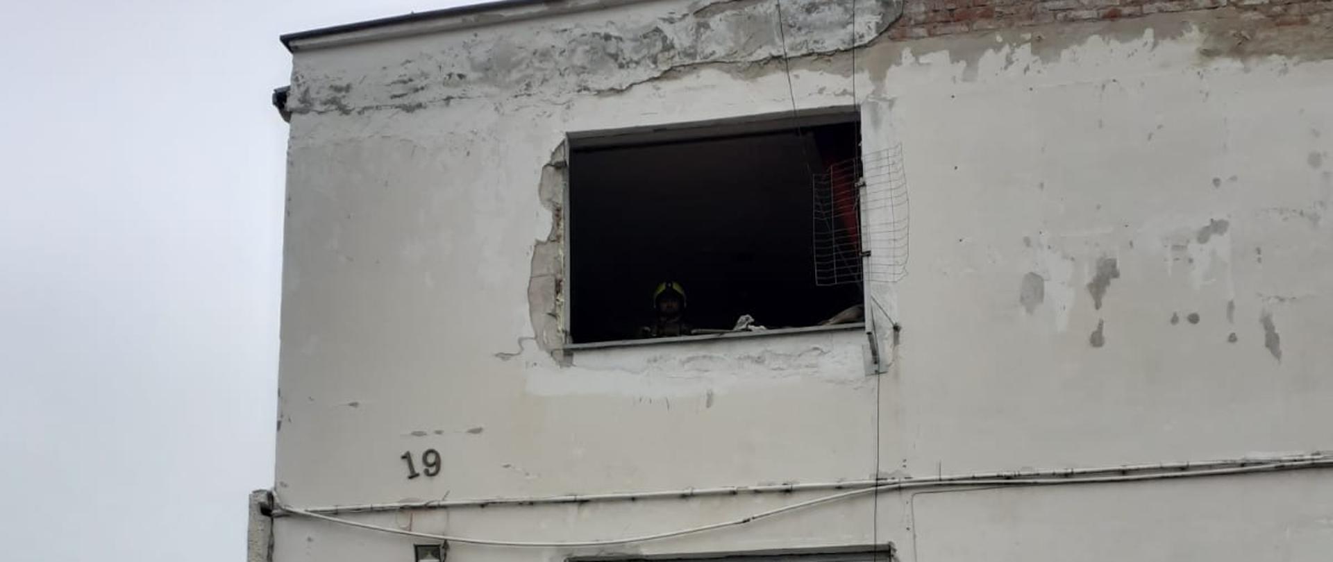 Fotografia przedstawia budynek koloru białego z uszkodzoną futryną okienną w wyniku wybuchu gazu. Na ziemi przed budynkiem leży wysadzone okno.