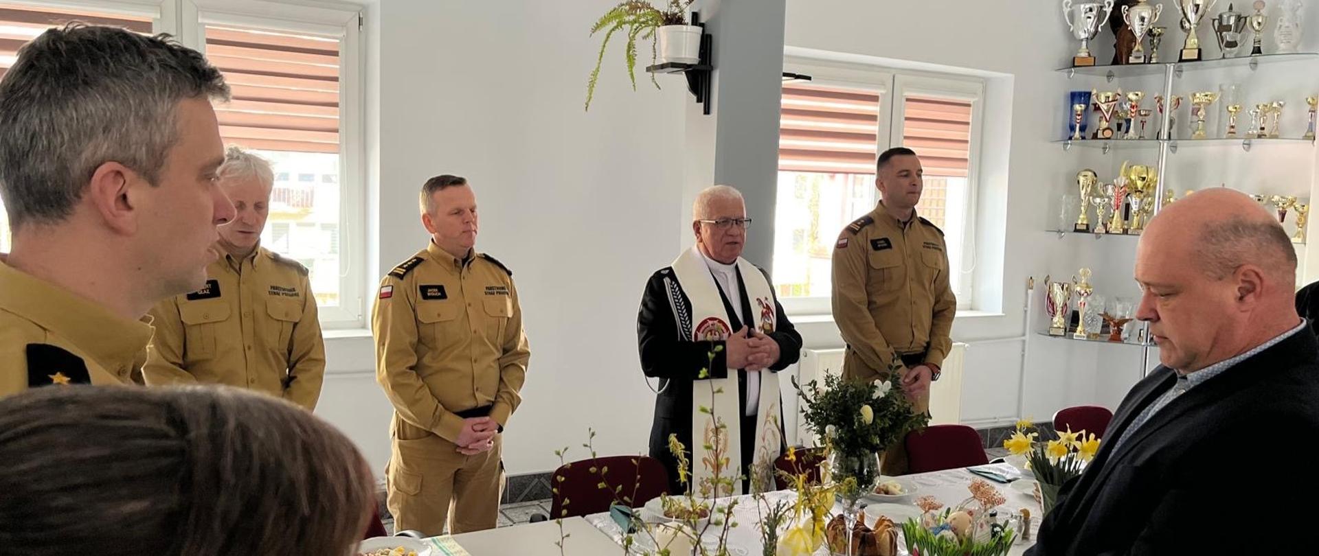 W pomieszczeniu strażacy uczestniczą w modlitwie, którą prowadzi ksiądz kapelan.