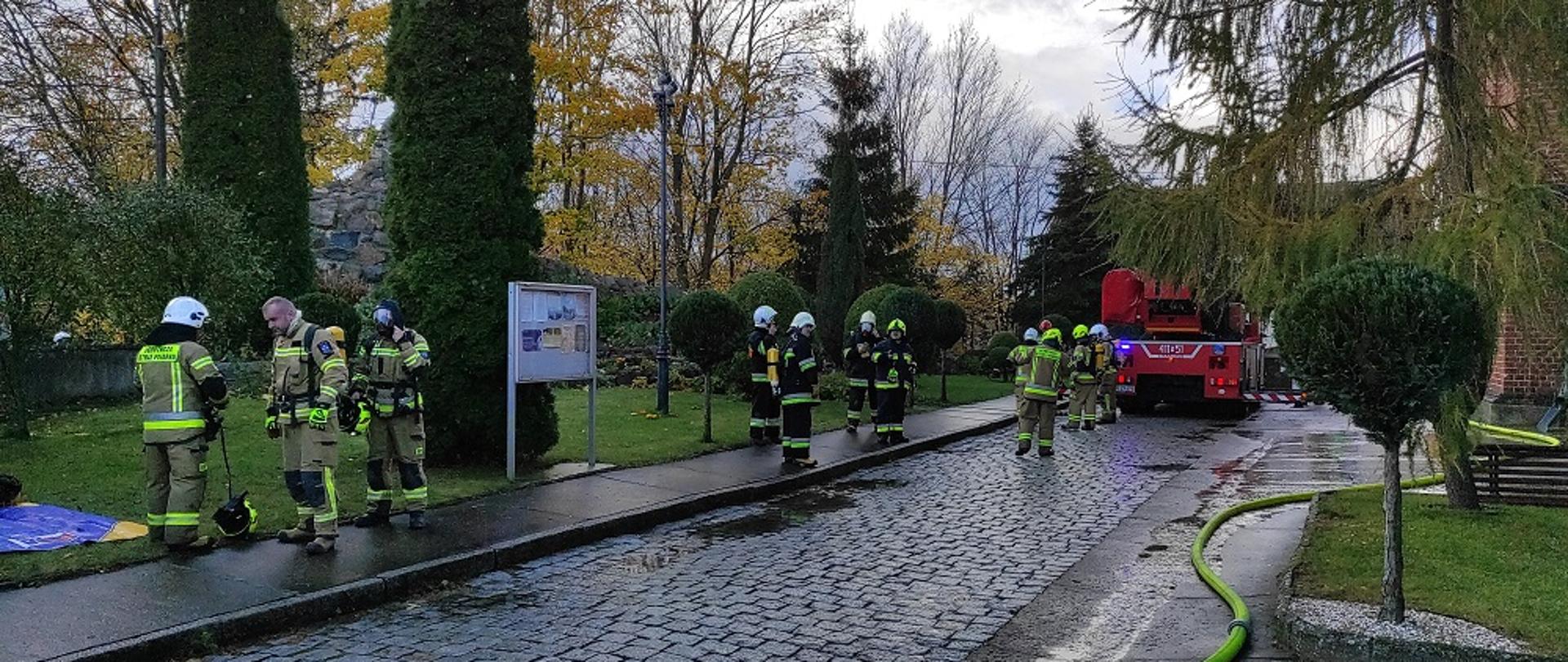 Przy kościele stoi samochód strażacki. Strażacy na chodniku przygotowują się do ćwiczeń.