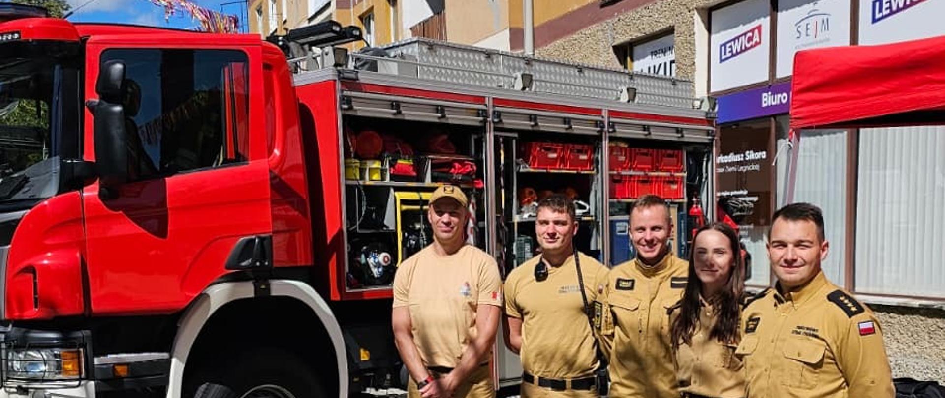 Na zdjęciu znajduje się grupa pięciu strażaków – czterech mężczyzn i jedna kobieta – pozująca z uśmiechem w słoneczny dzień na tle czerwono-białego wozu strażackiego marki Scania. Strażacy mają na sobie, piaskowe mundury służbowe z polskimi flagami i naszywkami na ramionach. Pojazd parkuje na kostce brukowej i ma otwarte boczne skrytki, w których widać specjalistyczny sprzęt gaśniczy oraz ratowniczy. Cała scena rozgrywa się na placu przed wielopiętrowym budynkiem mieszkalno-usługowym, a obok strażaków rozstawiono czerwony namiot promocyjny.