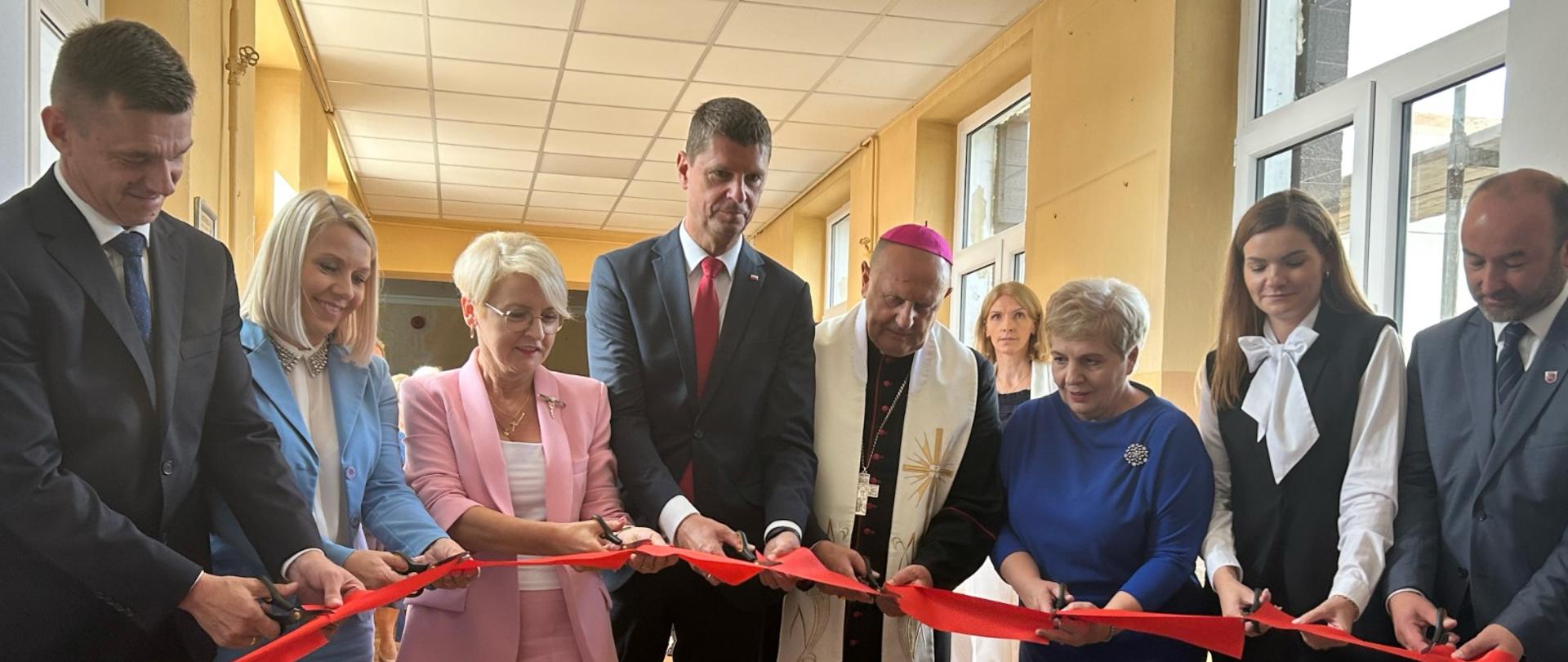 Wiceminister Piontkowski w otoczeniu kilku osób stoi w korytarzu szkoły i przecina czerwoną wstęgę.