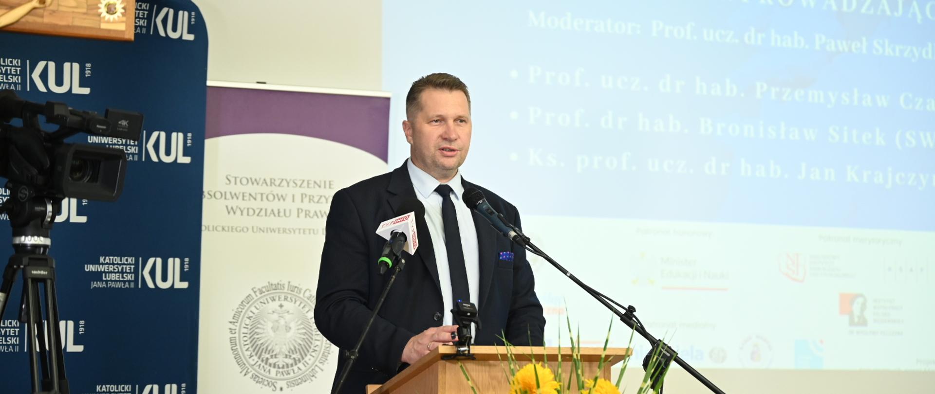 Minister Czarnek stoi za drewnianą mównicą i mówi do dwóch mikrofonów, przed mównicą bukiet żółtych kwiatów, za nim ścianka z napisem KUL.
