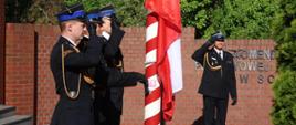 Na zdjęciu widzimy uroczyste podniesienie flagi państwowej, najprawdopodobniej z okazji Dnia Flagi Rzeczypospolitej Polskiej. Ceremonia odbywa się przed siedzibą Komendy Miejskiej Państwowej Straży Pożarnej w Sosnowcu.
Na pierwszym planie trzech strażaków w galowych mundurach, podczas gdy flaga Polski jest wciągana na biało-czerwony maszt. Jeden ze strażaków, znajdujący się po prawej stronie kadru, również salutuje, stojąc w oddaleniu od masztu. W tle widoczna jest czerwona ceglana ściana oraz zieleń drzew.