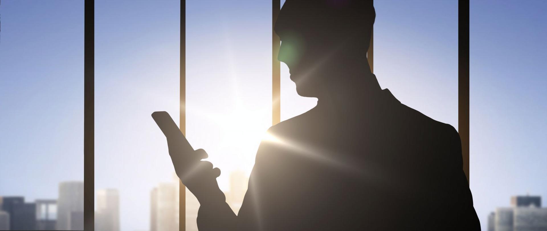 Osoba w cieniu z telefonem komórkowym, na tle okna.