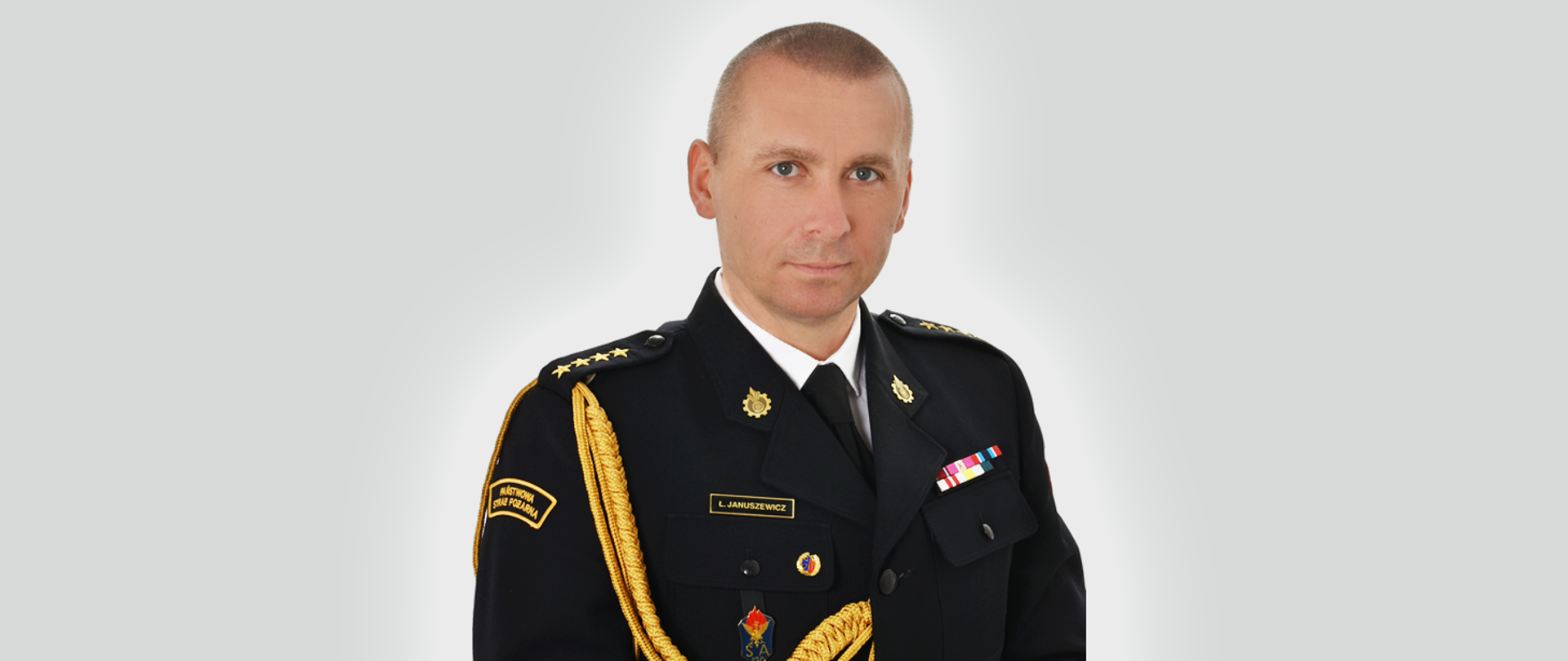 Zdjęcie przedstawia starszego kapitana Łukasza Januszewicza Komendanta Miejskiego PSP w Zielonej Górze w mundurze na szarym tle. 