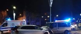Pora nocna. Na zdjęciu widać z lewej strony biały pojazd osobowy zderzony czołowo z szarym busem osobowym. W tle widać karetkę pogotowia i niebieskie światło sygnalizacyjne ze stojącego z prawej strony kadru pojazdu gaśniczego PSP.