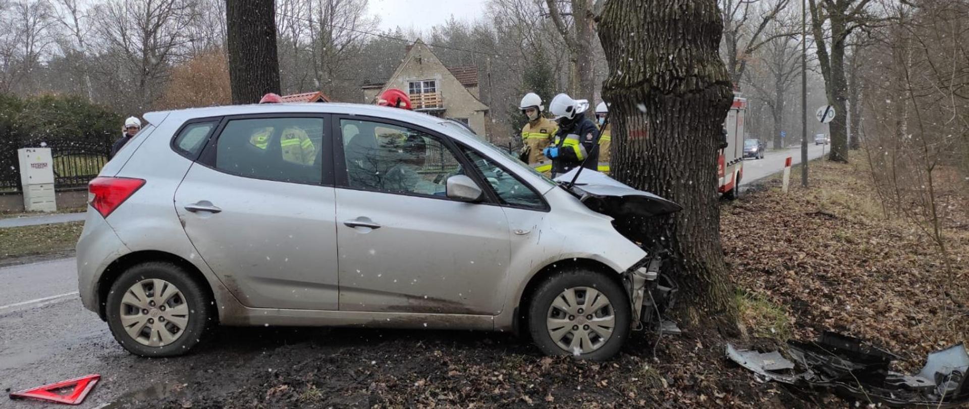 Samochód osobowy uderzył przodem w drzewo. Kierujący pojazdem znajduje się wewnątrz samochodu. Samochód częściowo stoi na jezdni i poboczu drogi.