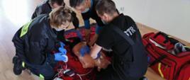 Zdjęcie przedstawia strażaków ćwiczących kwalifikowaną pierwszą pomoc związaną z nagłym zatrzymaniem krążenia.
W tle pomieszczenie świetlicy.
