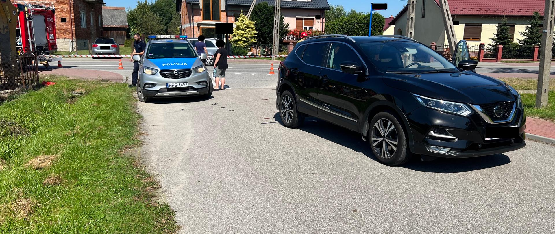 Na 1 planie samochód osobowy czarny biorący udział w zdarzeniu stojący przodem na jezdni, za nim na lewo radiowóz policyjny, w tle na lewo tył samochodu pożarniczego, w tle na wprost i na prawo domy jednorodzinne. W tle za samochodami widać strażaka PSP oraz Policjanta