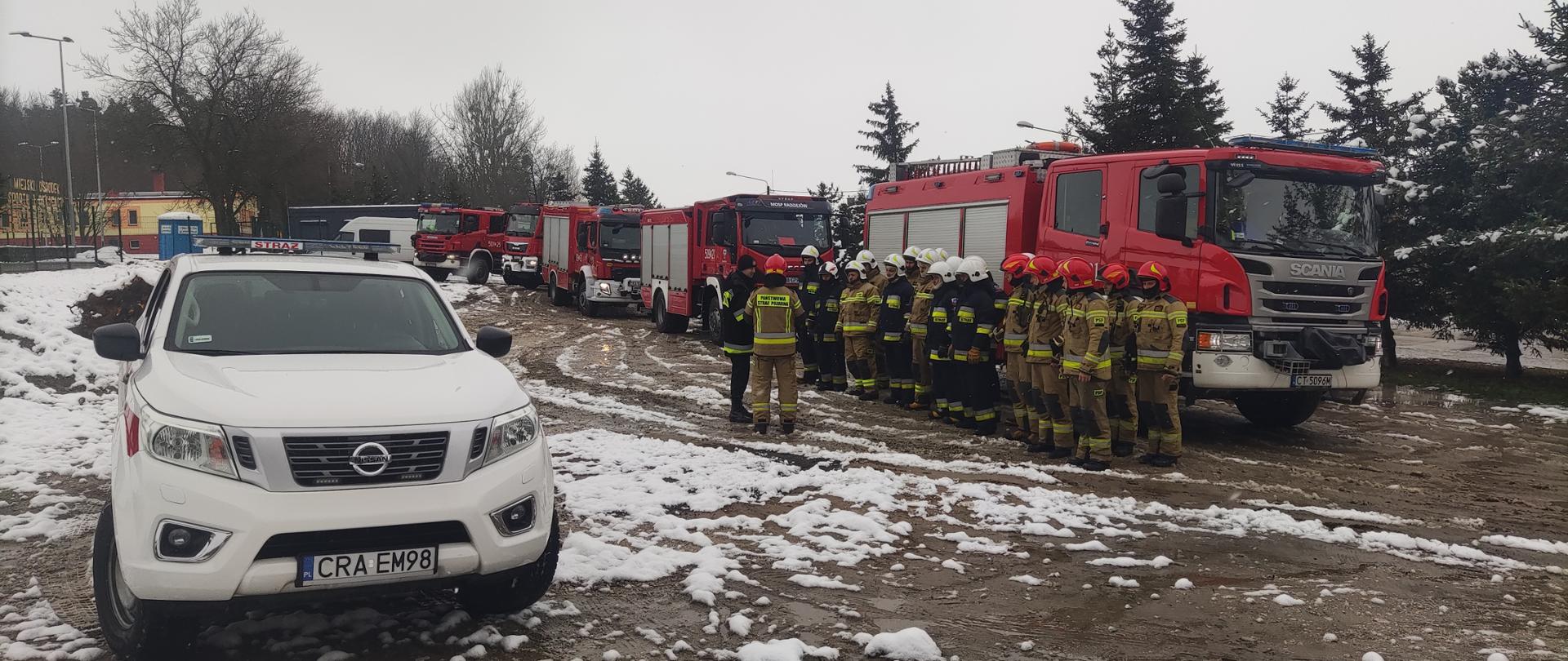 Zdjęcie na pierwszym planie przedstawia samochód operacyjny straży pożarnej koloru białego, na drugim planie strażaków PSP oraz OSP podczas odprawy z dowódcą JRG, na trzecim planie wozy strażackie koloru czerwonego