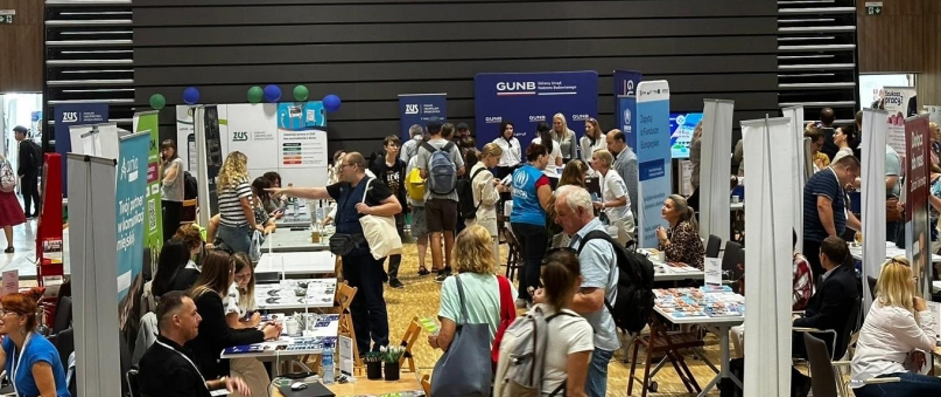 Wnętrze hali z licznymi stoiskami informacyjnymi i uczestnikami wydarzenia, widoczne banery ZUS i GUNB.