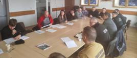 Uczestnicy narady siedzą przy dużym stole konferencyjnym podczas spotkania roboczego. Wśród obecnych znajdują się funkcjonariusze Państwowej Straży Pożarnej oraz przedstawiciele innych służb i instytucji, którzy analizują dokumenty i prowadzą dyskusję.