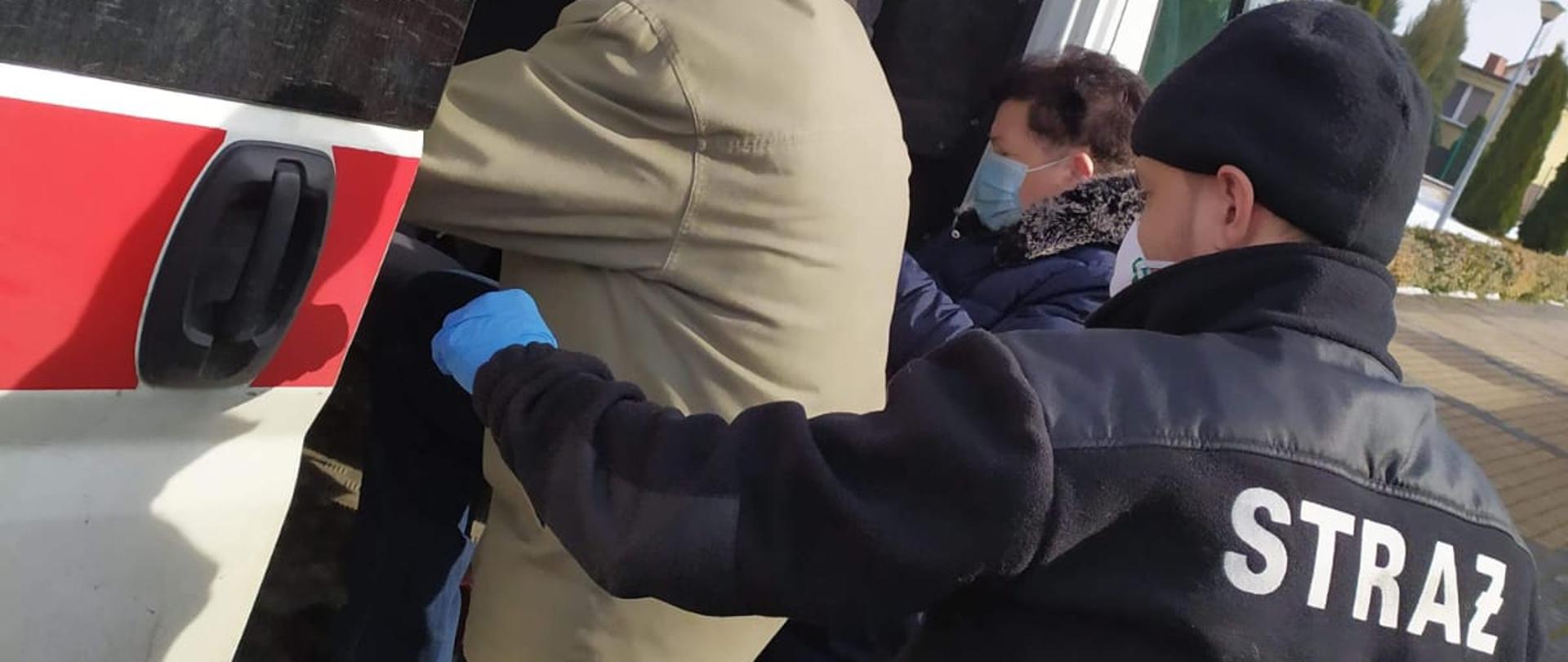 Zdjęcie przedstawia srebrny, strażacki samochód typu bus widziany z boku, do którego przez otwarte drzwi przesuwne wsiada mężczyzna w jasnej kurtce i niebieskich spodniach. Pomaga mu kobieta stojąca obok i strażak stojący za nim. Wszyscy mają usta zasłonięte maseczkami. 