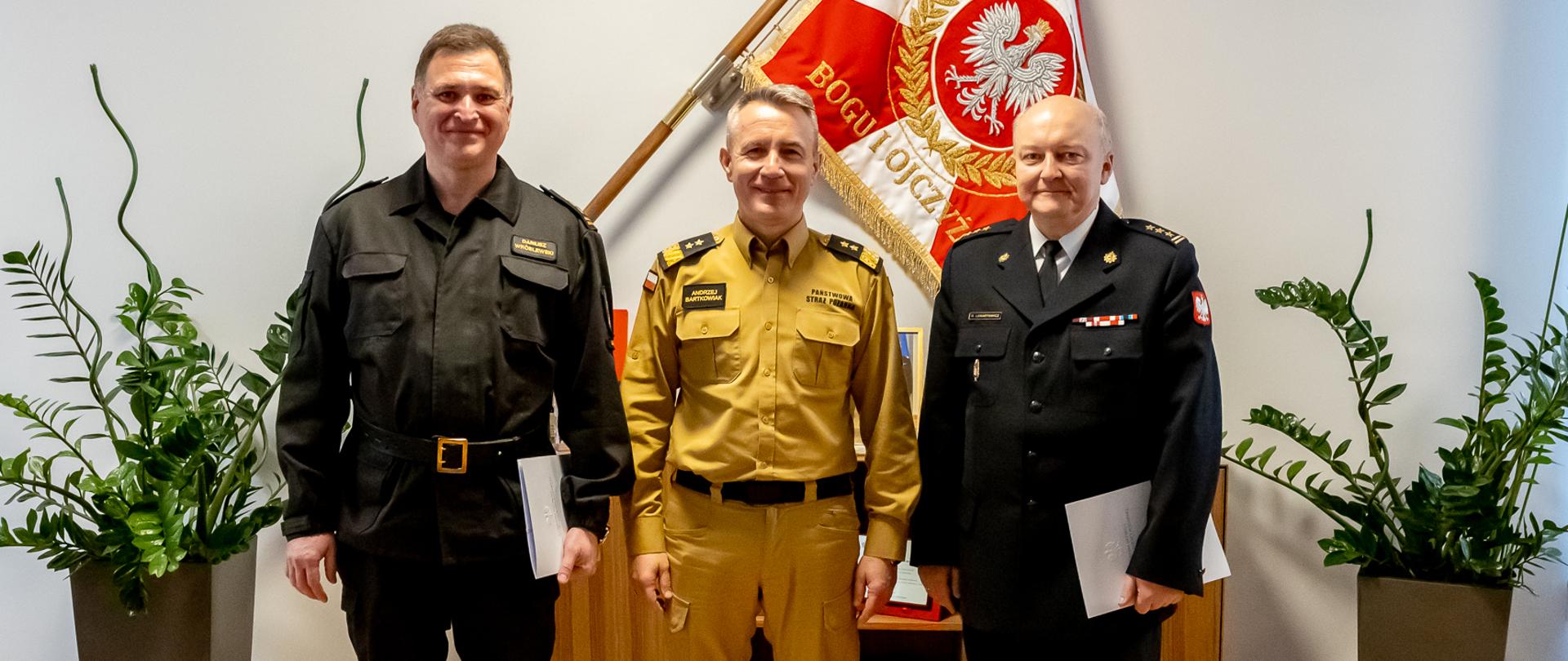 Na zdjęciu widać trzech strażaków. W środku stoi komendant główny PSP, w tle sztandar z godłem RP.